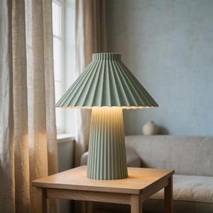 May include: A sage green table lamp featuring a wide, pleated conical shade and a matching cylindrical base with vertical fluting. The lamp emits a warm, soft glow from beneath the shade, illuminating the top of the wooden table surface.