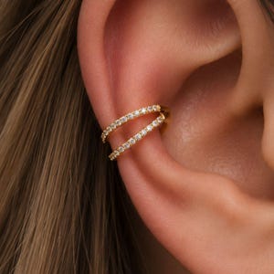 Peut inclure: Un ear cuff doré avec deux rangées parallèles de petits pierres précieuses transparentes. Le bijou est porté sur l'oreille, mettant en valeur le design et l'éclat des pierres. Un accessoire moderne.