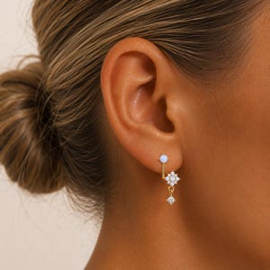 May include: A pair of gold earrings with a delicate design. One earring features a small, light blue orb at the earlobe, with a dangling floral cluster and a small, square-cut clear stone. The other earring has a matching light blue orb.