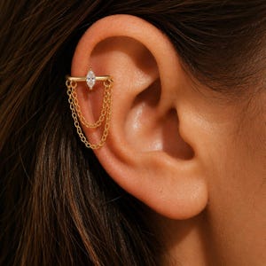 Peut inclure: Manchette d'oreille dorée ornée d'une pierre précieuse transparente en forme de marquise et de deux fines chaînes pendantes. La manchette d'oreille s'enroule autour du cartilage de l'oreille.