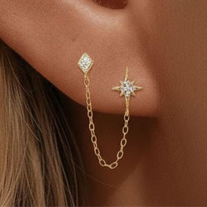 Peut inclure: Une boucle d'oreille double en or reliée par une chaîne délicate. Un clou est un motif en forme de losange incrusté de petites pierres claires, et l'autre est une étoile à quatre branches avec un groupe de pierres claires en son centre.