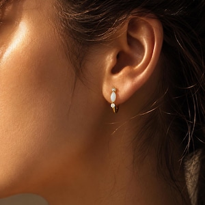 Peut inclure: Boucle d'oreille dorée avec une pierre blanche de forme ovale et deux petites pierres transparentes. La boucle d'oreille est portée sur l'oreille. Ce bijou délicat est un petit accessoire élégant.