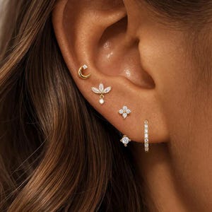 May include: A collection of gold-toned earrings, including a crescent moon stud, a floral design with a dangling pearl, a four-petal flower, a square stud, and a small hoop. The earrings are adorned with small, sparkling crystals.