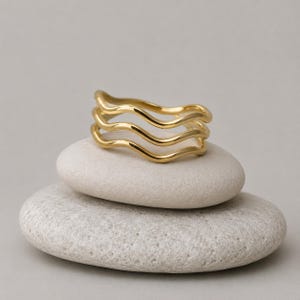 Op de afbeelding: Een goudkleurige ring bestaande uit drie met elkaar verbonden golvende banden die een gelaagd, vloeiend uiterlijk creëren. Het metaal heeft een gladde, gepolijste afwerking die het licht reflecteert en de continue, golvende lijnen benadrukt.