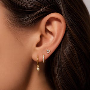Peut inclure: Gros plan sur une oreille ornée de plusieurs boucles d'oreilles dorées. Les boucles d'oreilles comprennent un petit cerceau avec un charm en forme de diamant pendant, un petit cerceau avec des cristaux incrustés et une puce avec trois petits cristaux.