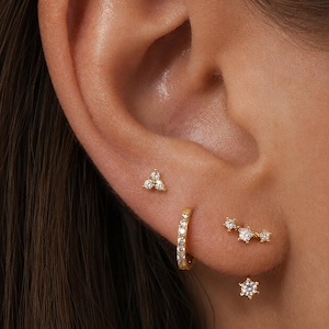 Op de afbeelding: Een set goudkleurige oorbellen met een kleine knop met drie stenen, een oorring bezet met heldere steentjes, een gebogen knop met drie stenen en een enkele solitaire knop met zes pootjes, gedragen in meerdere gaatjes.