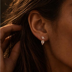 May include: A gold-toned earring set featuring two distinct stones: a small marquise-cut clear gemstone positioned at the top and a larger pear-shaped clear gemstone hanging directly below it, both secured in a metallic setting.