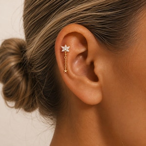 Könnte beinhalten: Ein goldfarbener Ohrschmuck mit einem blütenförmigen Stecker, besetzt mit klaren Edelsteinen. Von der Blüte hängt eine zarte Kette herab, die in einem kleinen Sternanhänger endet. Der Ohrring wird am oberen Ohr getragen und zeigt ein minimalistisches und elegantes Design.