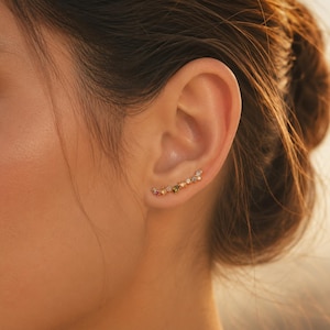 May include: A gold ear climber earring with a series of colourful gemstones. The gemstones include pink, green, yellow, and clear stones, arranged in a curved line along the ear. The earring is designed to be worn along the earlobe.