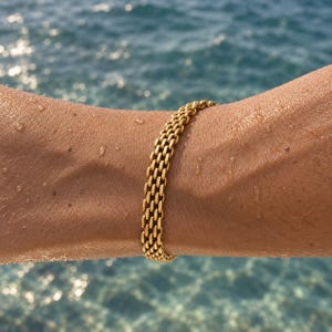 May include: A gold-toned bracelet featuring a dense, interlocking chain-link design. The bracelet is composed of multiple rows of small, uniform rectangular links that create a textured, woven appearance, resting against a sun-kissed skin surface.