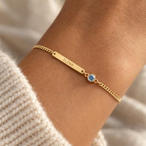 Peut inclure: Un bracelet à chaîne doré doté d'une barrette rectangulaire gravée du nom ELENA et d'une pierre précieuse ronde bleu clair sertie dans une monture. Les maillons métalliques délicats relient la barrette centrale et la pierre.