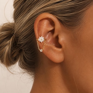 May include: A gold-colored ear cuff featuring a floral design with small, clear gemstones. A delicate chain connects the top floral piece to a smaller flower. The jewelry is worn on the ear.