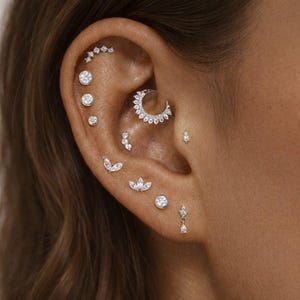Peut inclure: Une collection de piercings d'oreille argentés comprenant plusieurs clous à pierres transparentes, un anneau de daith en forme de croissant de lune, des motifs en forme de feuilles et une petite boucle d'oreille pendante sur le lobe.