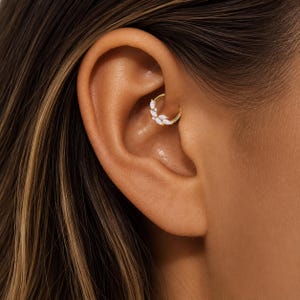 Peut inclure: Gros plan sur une boucle d'oreille dorée avec des accents de pierres précieuses blanches. La boucle d'oreille est placée dans le cartilage supérieur de l'oreille. Le teint est clair avec des reflets et des ombres.