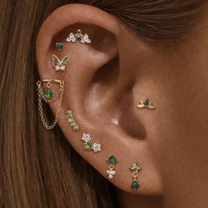 Peut inclure: Une collection de boucles d'oreilles dorées ornées de pierres vertes et de cristaux transparents. L'ensemble comprend un clou papillon, une manchette à chaînettes, des clous en forme de fleurs et divers motifs géométriques sur l'oreille.