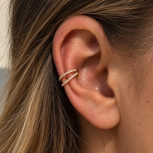 May include: A gold-toned ear cuff featuring two parallel bands encrusted with small, clear, sparkling stones. The jewelry piece wraps around the cartilage of the ear, creating a layered appearance with a polished metallic finish and bright accents.