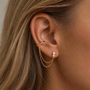 Peut inclure: Ensemble de boucles d'oreilles et de manchette d'oreille dorées. La manchette d'oreille s'enroule autour du haut de l'oreille, reliée par de fines chaînes à une boucle d'oreille à clous avec deux pierres précieuses rondes et claires. Le bijou est porté sur une oreille.