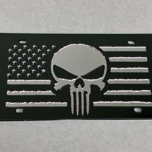 May include: Black license plate featuring a silver skull design over a stylized American flag. The skull is centered, with the flag's stars and stripes forming the background. The plate has four mounting holes.