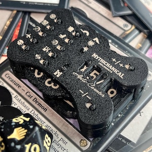 May include: A black, textured, bone-shaped mechanical counter for tabletop games. The device features the text "MYTHICHANICAL IMPLEMENTS" and "+/- Counter" with a numerical display showing "5" and "9". It includes various small engraved icons.