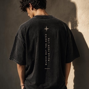 Puede incluir: Camiseta negra de gran tamaño con el texto "BLOCK OUT THE NOISE BUILD YOUR LIFE" en una línea vertical. Un diseño de rosa de los vientos está encima del texto. La camiseta tiene un aspecto vintage y lavado.