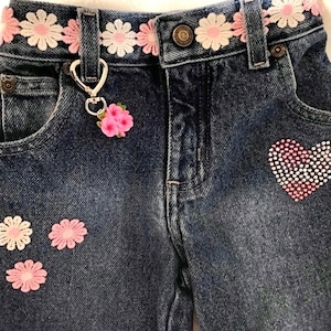 Pode incluir: Close-up de jeans azuis com cinto floral decorativo e detalhes de flores cor-de-rosa. Um design de coração de strass está no bolso direito. Um berloque prateado com flores cor-de-rosa pende da presilha.