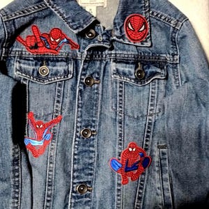May include: A blue denim jacket featuring a pointed collar, metal button closures, and four embroidered Spider-Man patches in red and blue. The interior label reads Place Est. 1989. The jacket includes two chest pockets and long sleeves.
