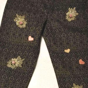 May include: Dark brown patterned pants with gold and pink embellishments. The pants feature a decorative waistband, floral lace appliques, and heart-shaped patches. The fabric has a repeating geometric design.