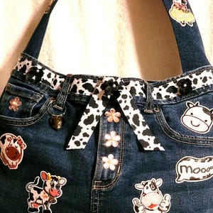 May include: Denim handbag with cow-themed patches and accents. The bag features a dark blue denim base, adorned with embroidered cow patches, a black and white cow print trim, and a decorative bow. The straps are also denim with cow-themed embellishments.