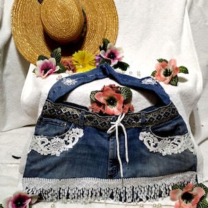 May include: A blue denim bag featuring a wide strap, a decorative patterned waistband, and white lace trim along the pockets and bottom edge. The bag includes a white drawstring closure and is adorned with white floral lace appliques.