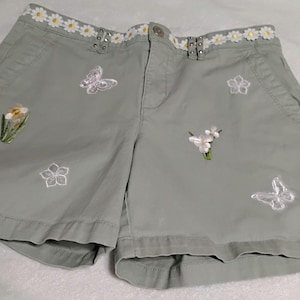 May include: Sage green shorts with embroidered butterflies and flowers. The waistband features white daisy trim. The shorts have side pockets and button details. A spring-themed garment.