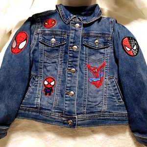May include: A blue denim jacket featuring a classic collar, metal button closures, and two chest pockets. The jacket is decorated with multiple embroidered patches of the character Spider-Man, including various mask designs and a full-body pose.