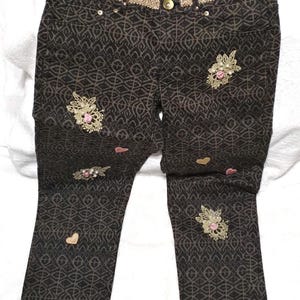 May include: Dark brown pants featuring a repeating geometric pattern, a gold-toned metallic waistband trim, and several floral lace appliqués with small pink rose accents and scattered heart-shaped embellishments across the front legs.