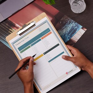 May include: A wooden clipboard holds a "Vacation Budget Tracker" form with colorful sections. A black pen is poised over the form, ready to write. A laptop, plant, and glass are in the background, suggesting a workspace.