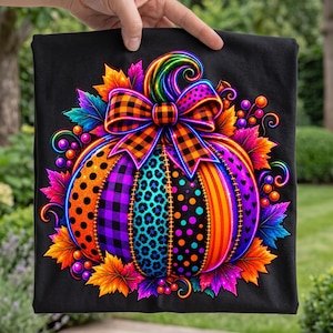 Könnte beinhalten: Ein schwarzes T-Shirt mit einer bunten Kürbisgrafik. Der Kürbis ist mit verschiedenen Mustern wie Leopardenmuster, Punkten und Karos verziert und wird von einer großen orange-schwarzen Karo-Schleife sowie herbstlichen Blättern gekrönt.