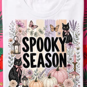 Könnte beinhalten: Ein weißes T-Shirt mit einem Grafikdruck und dem Text SPOOKY SEASON in Schwarz. Das Design zeigt zwei schwarze Katzen mit rosa Schleifen, Kürbisse, Laternen, Schmetterlinge und verschiedene Blumen in Pastellfarben vor einem gestreiften Hintergrund.