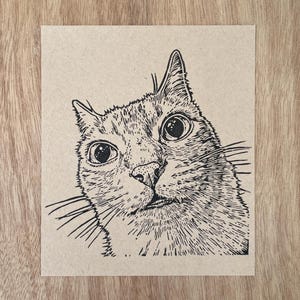 Puede incluir: Una impresión cuadrada sobre papel marrón claro que presenta una ilustración en tinta negra de una cabeza de gato. El gato está representado con ojos circulares grandes, bigotes prominentes y una textura de pelaje detallada.