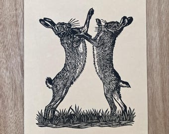 May include: A black ink block print on light beige paper featuring two hares standing on their hind legs and facing each other in a boxing stance. The hares are positioned above a patch of grass, with detailed fur textures and long ears.