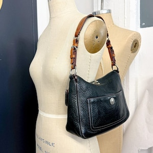 May include: A black pebbled leather shoulder bag featuring a tan leather strap with silver-toned hardware, a front exterior pocket with a silver circular emblem, and a top turn-lock closure. The bag has visible stitching and a hanging leather tag.