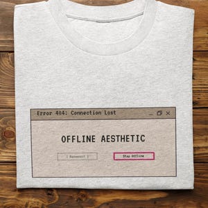 以下が含まれることがあります： ライトグレーのクルーネックTシャツ。コンピューターのエラーウィンドウのグラフィックプリントが施されています。ウィンドウには「Error 404: Connection Lost」、「OFFLINE AESTHETIC」というテキストと、ピンクの枠で囲まれた「Reconnect」および「Stay Offline」という2つのボタンが表示されています。