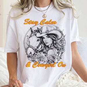 Può includere: T-shirt bianco con grafica di una donna con cappello da cowboy e un cavallo, circondati da rose. Il testo "Stay Calm & Cowgirl On" è in giallo e arancione. La maglietta ha maniche corte.