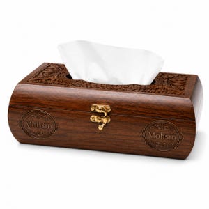 Könnte beinhalten: Ein rechteckiger Taschentuchbox-Halter aus dunkelbraunem Holz mit polierter Oberfläche. Die Oberseite weist aufwendige florale Schnitzmuster auf. Die Vorderseite enthält einen goldfarbenen Metallverschluss und zwei kreisförmige Gravuren mit dem Namen Mohsin.