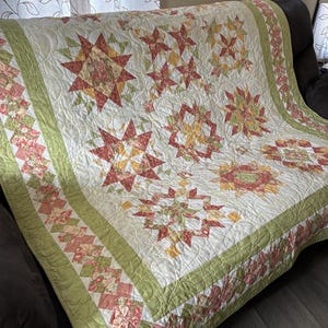 May include: A patchwork quilt featuring a repeating star pattern in shades of red, yellow, and green on a cream background. The quilt is bordered by a solid light green fabric and a decorative strip of small, multi-colored floral squares.