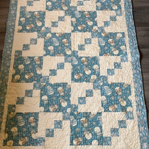 May include: A quilted blanket with a winter theme. The blanket features a pattern of white and blue squares, with a border of snowflakes. The blue squares are decorated with snowmen. The blanket is approximately 60 inches long and 40 inches wide.