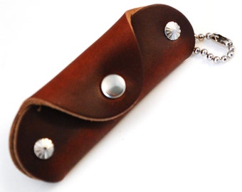 Könnte beinhalten: Ein brauner Leder-Schlüsselhalter mit einer silbernen Kugelkette und einem Verschluss. Der Schlüsselhalter ist mit drei silbernen Nieten befestigt. Das Leder hat eine satte, dunkelbraune Farbe.
