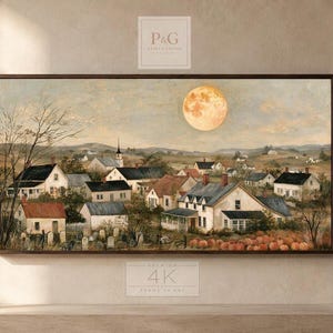 May include: A horizontal framed wall art print depicting a rural village scene under a large full moon. The landscape features multiple houses with pitched roofs, a church steeple, bare trees, and a field of orange pumpkins in the foreground.