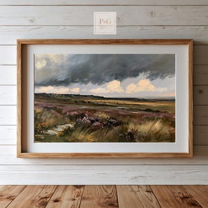 Könnte beinhalten: Ein gerahmtes Landschaftsgemälde, das eine weite Moorlandschaft unter einem dramatischen, dunklen und stürmischen Himmel zeigt. Im Vordergrund sind strukturierte Gräser und violette Heidekrautgewächse zu sehen, während am Horizont ferne Hügel liegen. Heller Holzrahmen.
