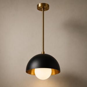 Könnte beinhalten: Eine Pendelleuchte mit einem mattschwarzen, kuppelförmigen Lampenschirm und einer metallisch goldenen Innenseite. Eine weiße, kugelförmige Glühbirne hängt an einer geraden, messingfarbenen Stange, die an einem runden Deckenbaldachin befestigt ist.