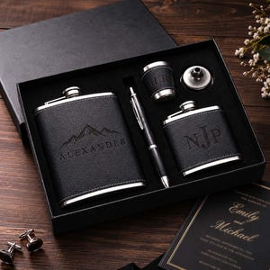 May include: A black gift box containing a large black leather-wrapped flask engraved with a mountain graphic and the name ALEXANDER, a smaller flask with the initials NJP, a pen, a small cup with initials HR, and a metal funnel, plus two cufflinks.
