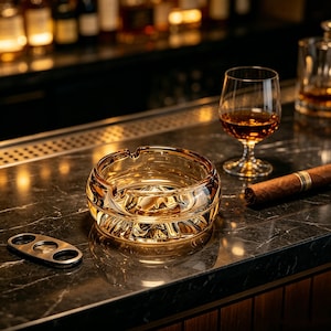 Op de afbeelding: Een heldere, amberkleurige glazen asbak met een sigaar, een glas amberkleurige vloeistof en een sigarenknipper op een donker aanrecht. De asbak heeft een cirkelvormig ontwerp met een gestructureerd interieur. Een bar met flessen staat op de achtergrond.