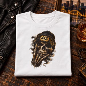 Op de afbeelding: Een wit t-shirt met een grafische print van een schedel die een zwarte muts draagt met het getal 412 op een gouden patch. De schedel is afgebeeld met skeletvingers bij het gezicht, omringd door wervelende grijze rook en druppels.